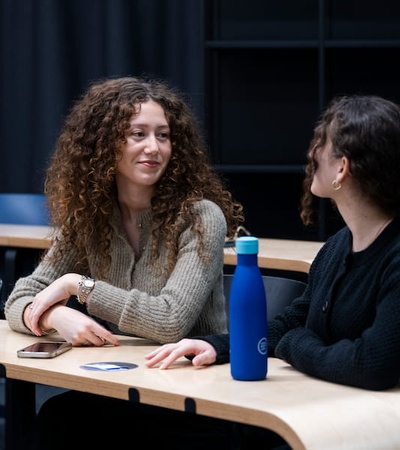 Two women with curly hair are having a conversation in a modern classroom.