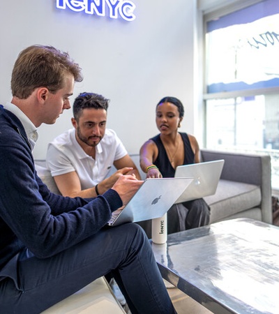 Three people are engaged in a discussion while working on laptops in a modern office setting.