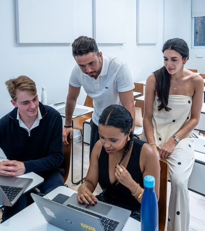 A group of four young adults collaborate around a laptop in a modern classroom setting.