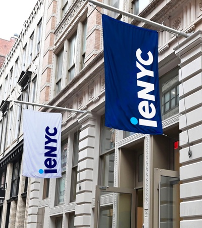 A view of a city street with buildings displaying blue 'ieNYC' flags.