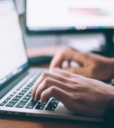 Person typing on a laptop with another monitor in the background.