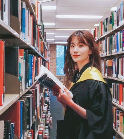 A graduate in a black robe holding a book in a library aisle, smiling at the camera.