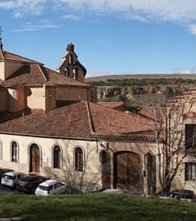 Panoramic view of an ancient monastery complex with terracotta roofs surrounded by sparse vegetation and a clear blue sky.