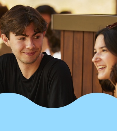 A young man and two women are smiling and chatting at an outdoor table.