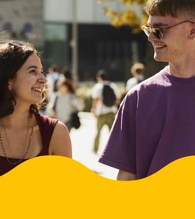 Two young adults smiling and talking to each other on a sunny day at an outdoor event.