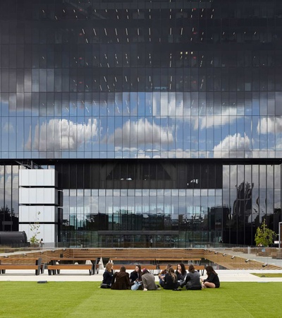 A group of people sitting on the grass in front of a modern glass building with a unique architectural design.