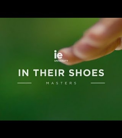 A promotional image featuring a close-up of a human finger pointing, with the text 'IE University - In Their Shoes, Masters' overlayed on a blurred green background.