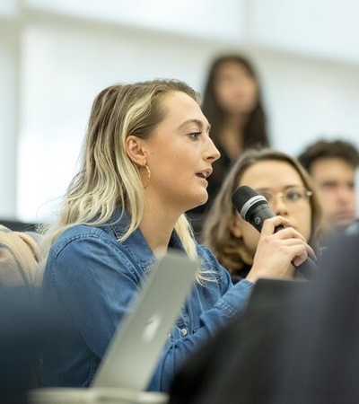 A woman holding a microphone speaks during a discussion in a classroom setting.