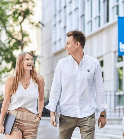 Two young adults walking and laughing together on a university campus, one of them holding books.