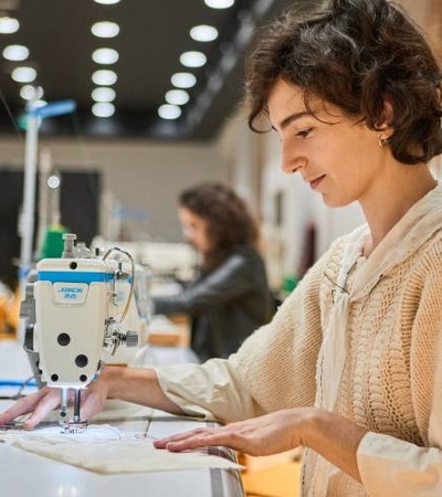 A woman is sewing fabrics using a machine in a well-lit workshop.