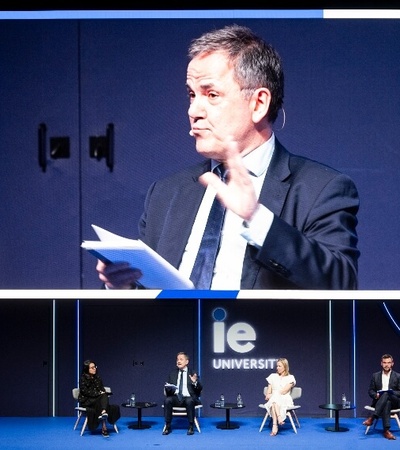 A man speaking on stage at an event with an audience and a large screen displaying his image in the background.