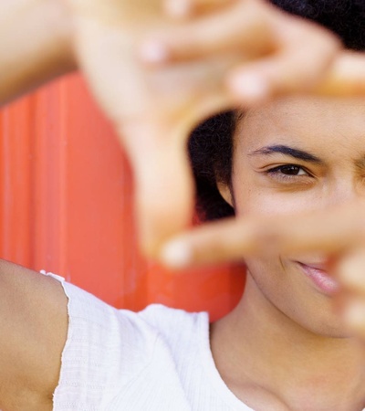 A young person with an afro smiling and making a frame with their hands in front of a red background.
