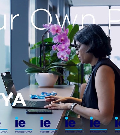 A woman working on a laptop at a modern desk with plants and flowers in the background.