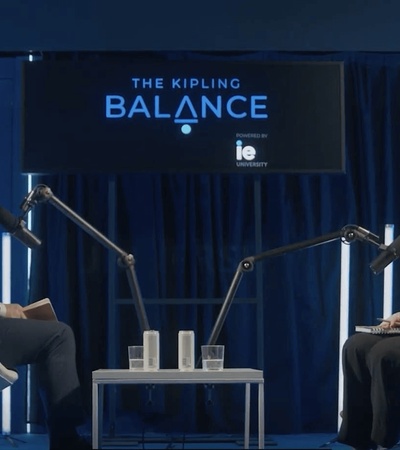 A discussion taking place on a stage with two speakers, seated in modern chairs, and surrounded by blue decor.