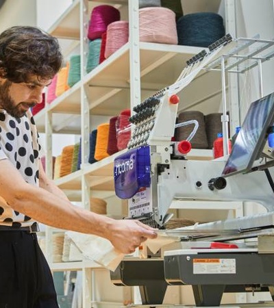 A man operating an embroidery machine in a textile workshop, surrounded by colorful thread spools.