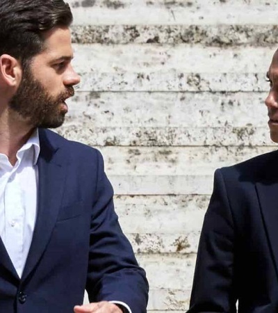 Two men in suits having a conversation against a textured wall.
