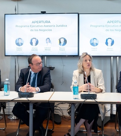 Four people sitting at a panel discussion table in a conference room, with projectors displaying the word 'APERTURA' in the background.