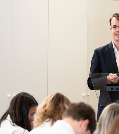 A young man giving a presentation at a podium in front of an audience of seated women.