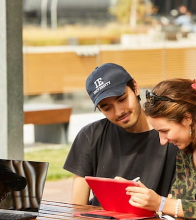 Two young adults, a male and a female, are looking at a red folder together at an outdoor table surrounded by others.