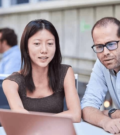 A group of colleagues, including a woman and two men, are engaged in a discussion around a laptop at an outdoor table.