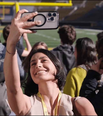 A woman is taking a selfie with her phone at a stadium event, smiling joyfully.