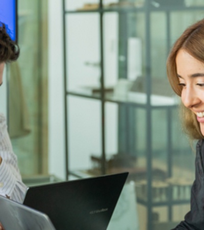 A man and a woman in business attire are discussing over a laptop in a modern office setting.