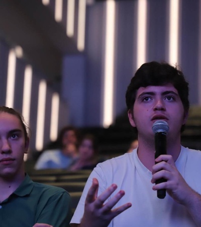 A young man speaks into a microphone while sitting next to a focused young woman in a lecture hall.