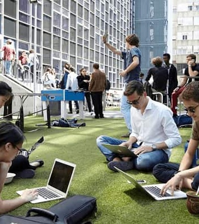 People using laptops while sitting on the grass in a busy outdoor city area.