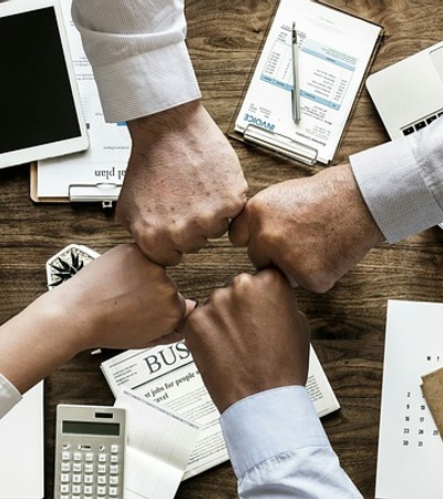 Four people fist bumping over a busy meeting table filled with electronic devices, documents, and coffee cups.