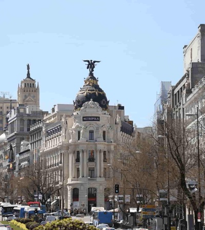 A busy urban street scene featuring classic architecture and a statue on top of a building, under a clear blue sky.