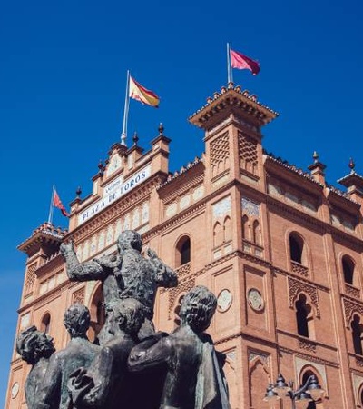 A view of a historic bullfighting arena with a bronze statue in the foreground and flags flying atop under a clear blue sky.