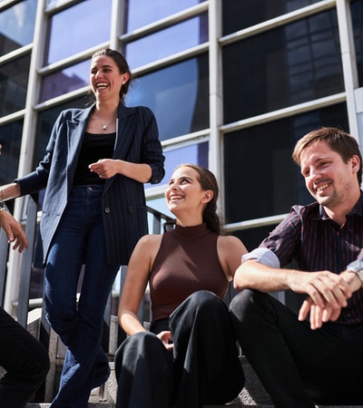Four people happily chatting and laughing outside a modern office building on a sunny day.