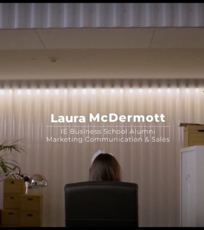 A woman sits at a desk in an office with a sign above reading 'Laura McDermott, IE Business School Alumni, Marketing Communication & Sales'.