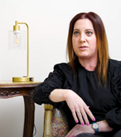 A woman with red hair sitting on a couch in a room beside an antique wooden table and a lamp.