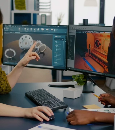 Two women working attentively at a dual-monitor computer setup, engaged in graphic design and discussing a 3D model.