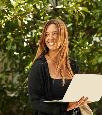 A young woman smiles while holding a laptop, standing outdoors surrounded by green foliage.
