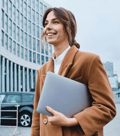 A smiling professional woman in a brown coat carries a laptop while walking outside a modern building.
