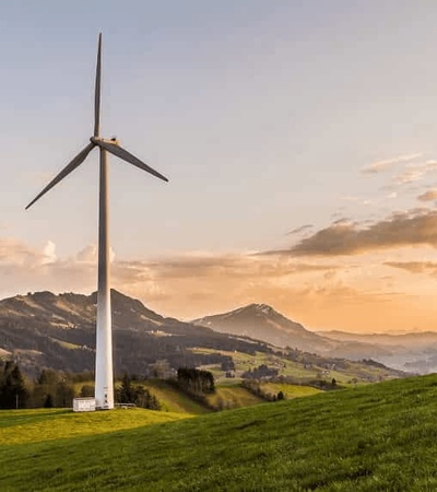A single wind turbine stands on a green hillside at sunset with mountains in the background.