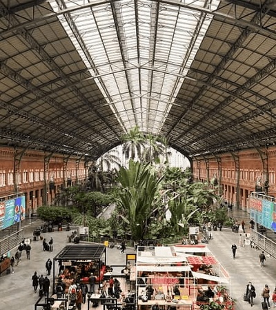 A spacious indoor train station featuring a large tropical garden, shops, and bustling commuters under a grand glass roof.