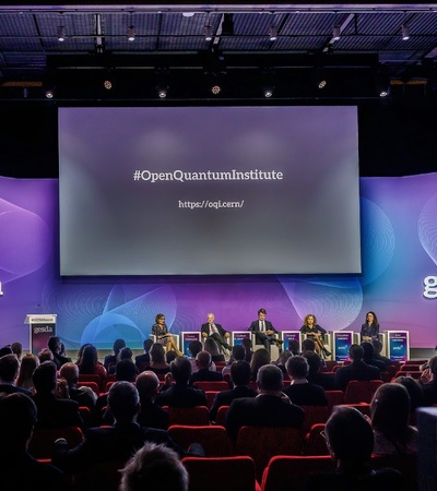 A conference hall with a panel of speakers, audience, and banners related to quantum institutes.