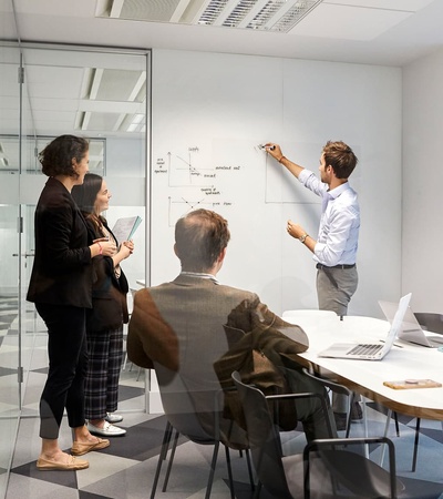 A team meeting in a modern office space with a person presenting on a whiteboard.
