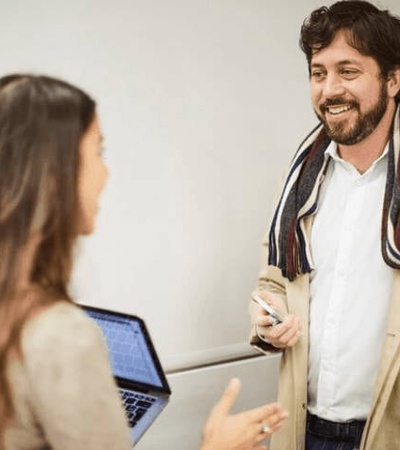 A man and a woman having a conversation in a classroom, with the man smiling and the woman holding a laptop.