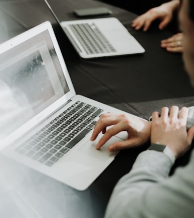 A person is working on a laptop at a table with another laptop in the background.