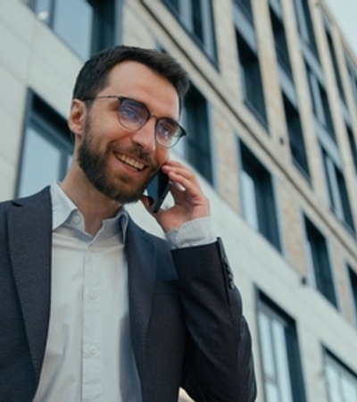 A man in a suit smiles while talking on his phone in front of a modern building.