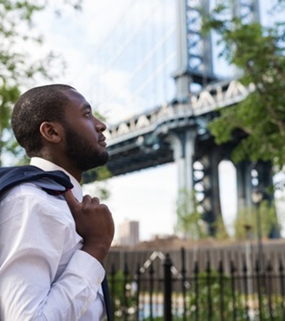 A man in a suit stands near a bridge, looking thoughtfully into the distance.