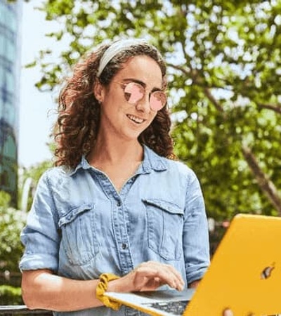 A young woman with curly hair wearing glasses and a denim shirt is smiling while using a yellow laptop outdoors in a city setting.