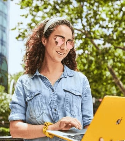 A woman with curly hair and glasses is using a yellow laptop outdoors in an urban setting.