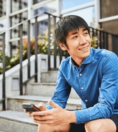 A young man sitting on steps outside a building, smiling while looking away and holding a smartphone.