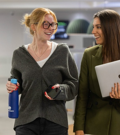 Two women are smiling and chatting while walking in a modern office environment.