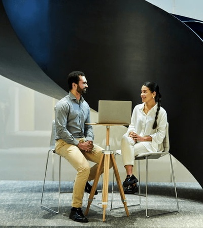A man and a woman sitting and talking under a large, spiral staircase in a modern building.